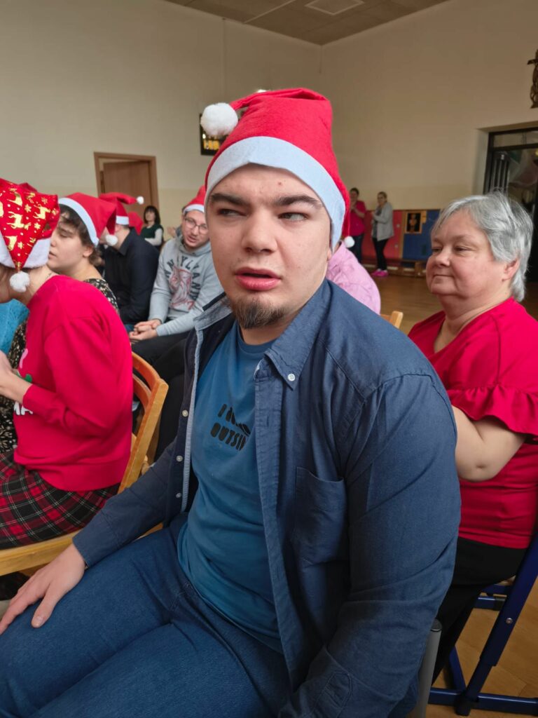 Grupa dzieci i dorosłych. Dzieci maja na głowach czapki mikołajowe. Wszyscy siedzą na sali i uważnie słuchają.