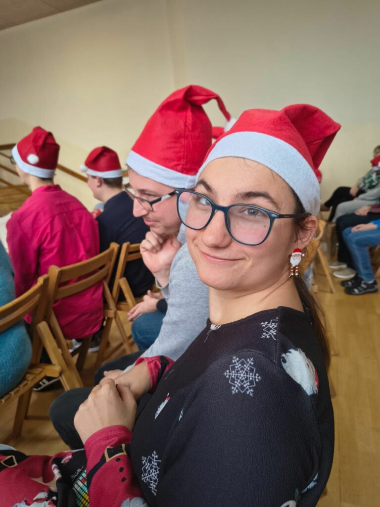 Uśmiechnięta młoda kobieta w okularach oraz mężczyzna w czapkach mikołaja. W tle pozostali uczestnicy spotkania.