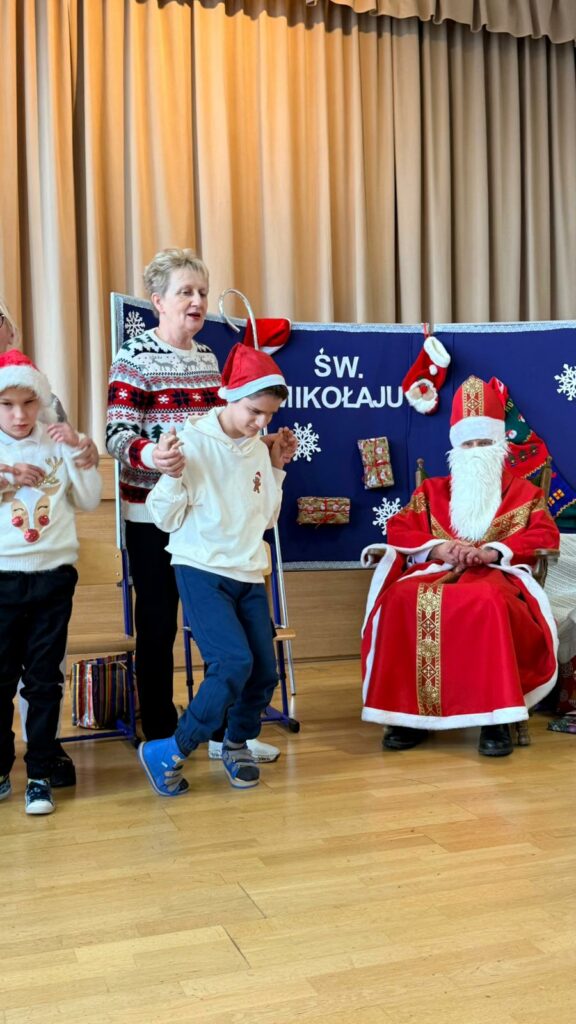 Chłopiec w czapce Mikołaja tańczy, prowadzony przez opiekunkę, a obok nich siedzi osoba przebrana za św. Mikołaja na tle ozdobionej sceny.