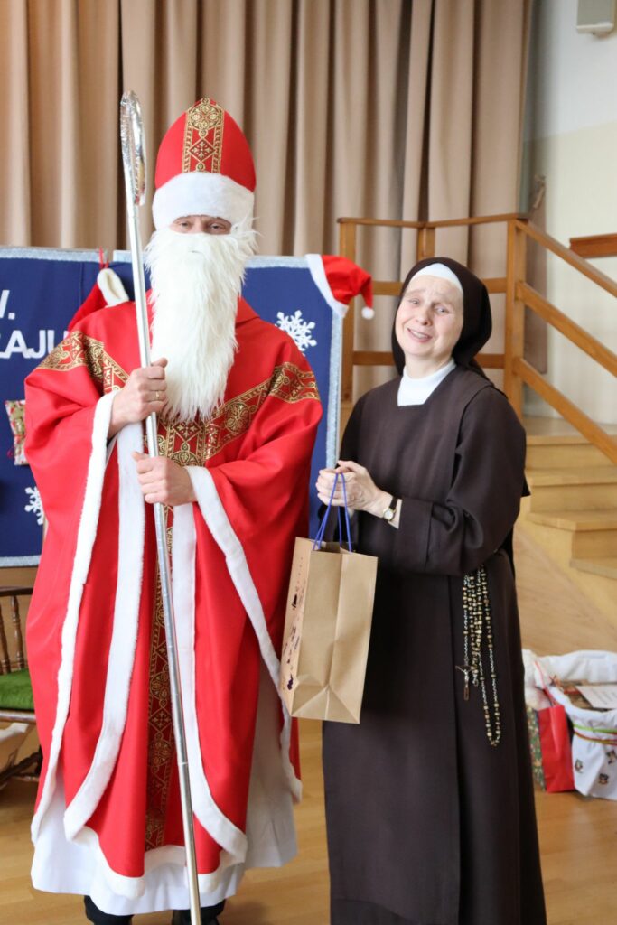 Św. Mikołaj w czerwonym stroju i siostra zakonna stoją obok siebie; siostra trzyma papierową torbę z prezentem i uśmiecha się w stronę aparatu.