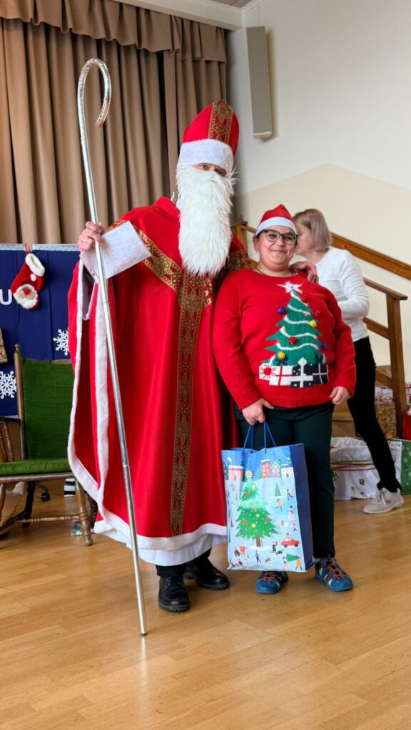 Św. Mikołaj w czerwonym stroju i Dziewczynka w czerwonym świątecznym swetrze stoją obok siebie; dziewczyna trzyma papierową torbę z prezentem i uśmiecha się w stronę aparatu.
