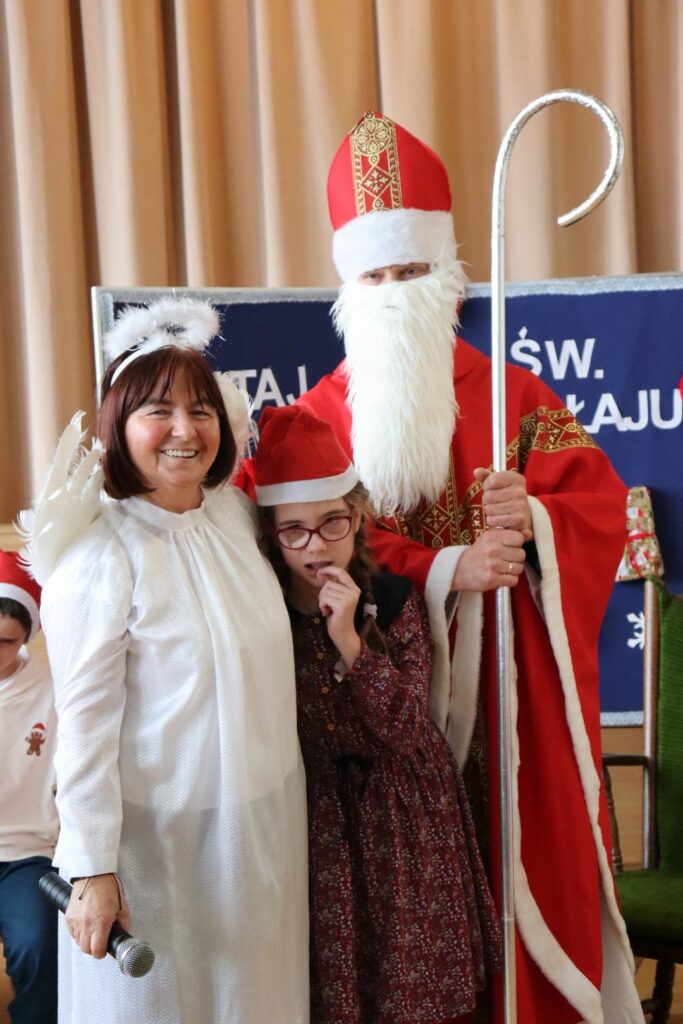 Kobieta przebrana za anioła, mężczyzna w stroju św. Mikołaja i dziewczynka w świątecznej czapce stoją razem, uśmiechając się.