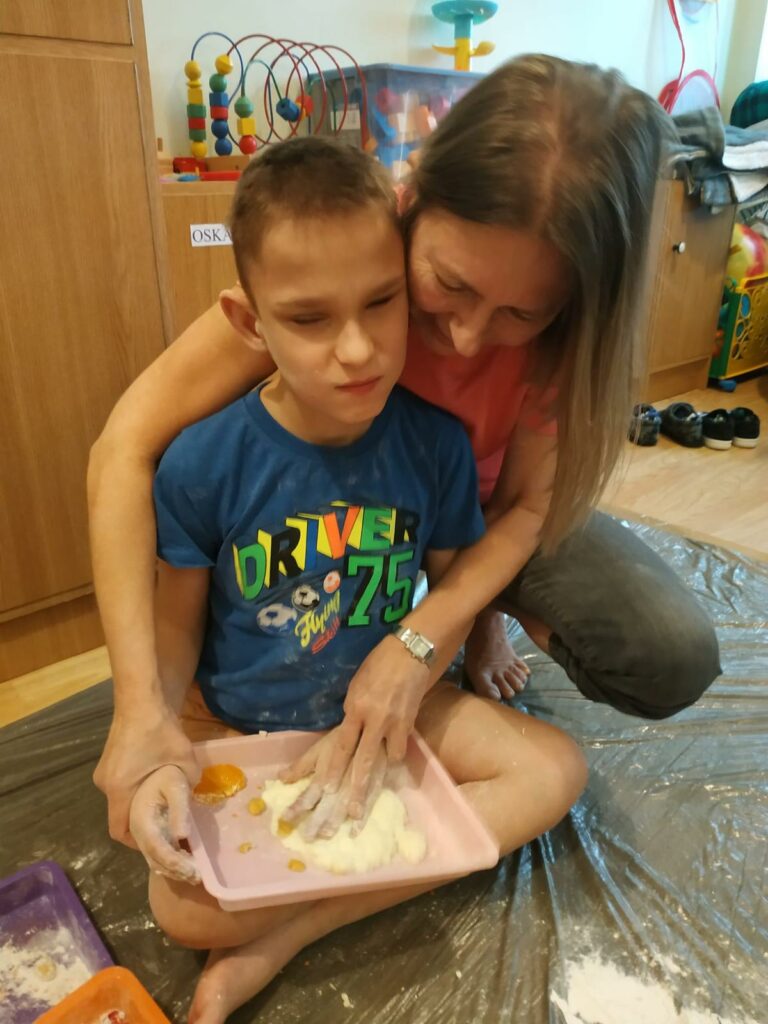 Chłopiec bawi się rękami w masie plastycznej, obok niego osoba dorosła wspiera goi pomaga w zabawie.