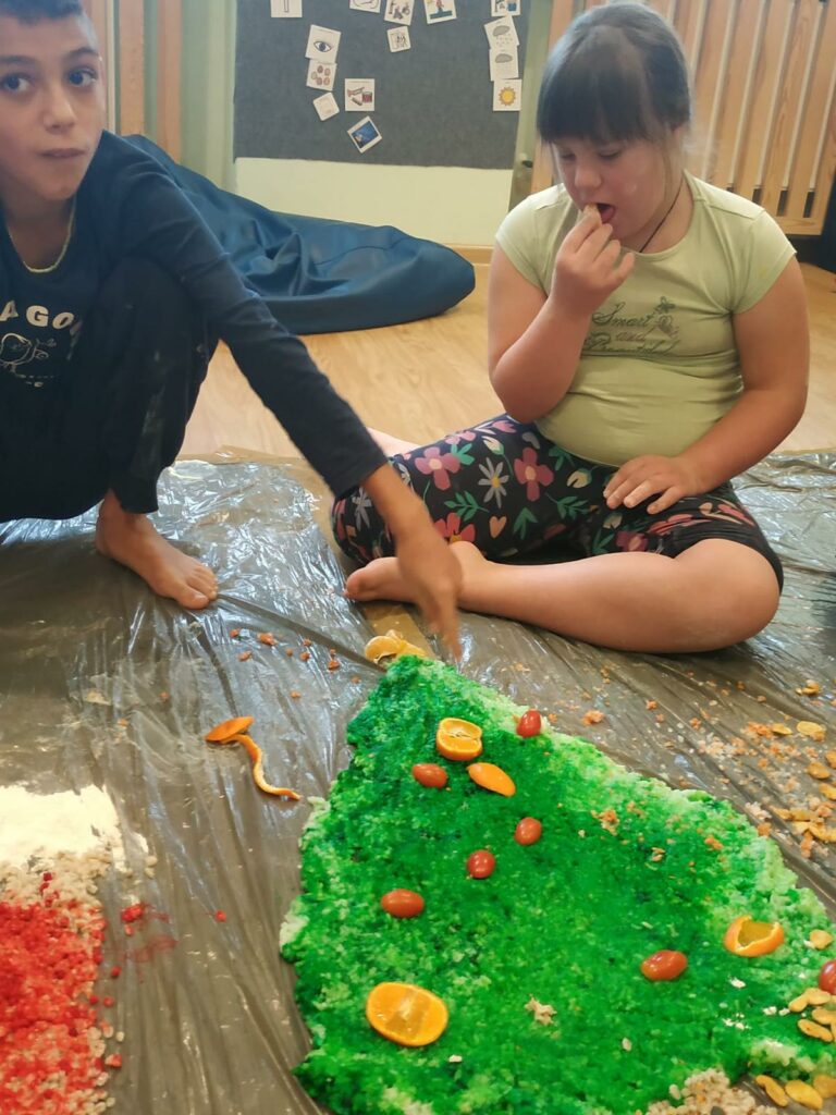 Dzieci tworzą kreatywną scenerię na podłodze, rozkładając kolorowy materiał sypki w kształt choinki