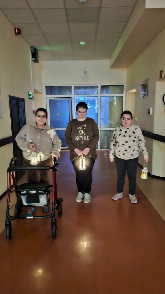 Trzy osoby idą korytarzem, niosąc zapalone lampiony; jedna osoba korzysta z chodzika.