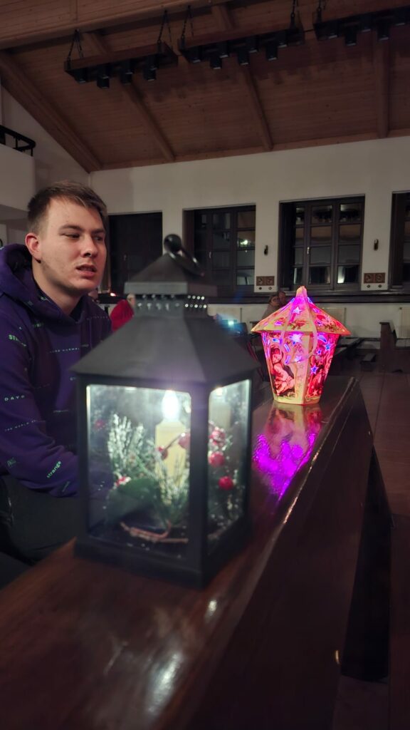 Młody mężczyzna siedzi w kościele przy ławce, obok zapalone dekoracyjne lampiony.