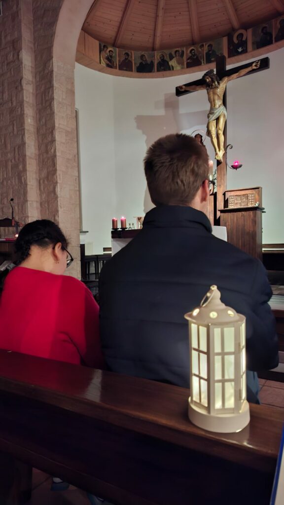 Wnętrze kościoła z krzyżem nad ołtarzem; wierni siedzą w ławkach, na pierwszym planie lampion.