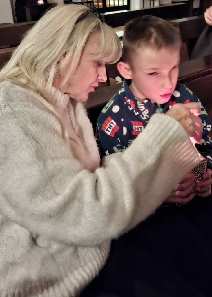 Starsza kobieta i chłopiec siedzą w ławce w kościele. Kobieta zapala chłopcu lampion.