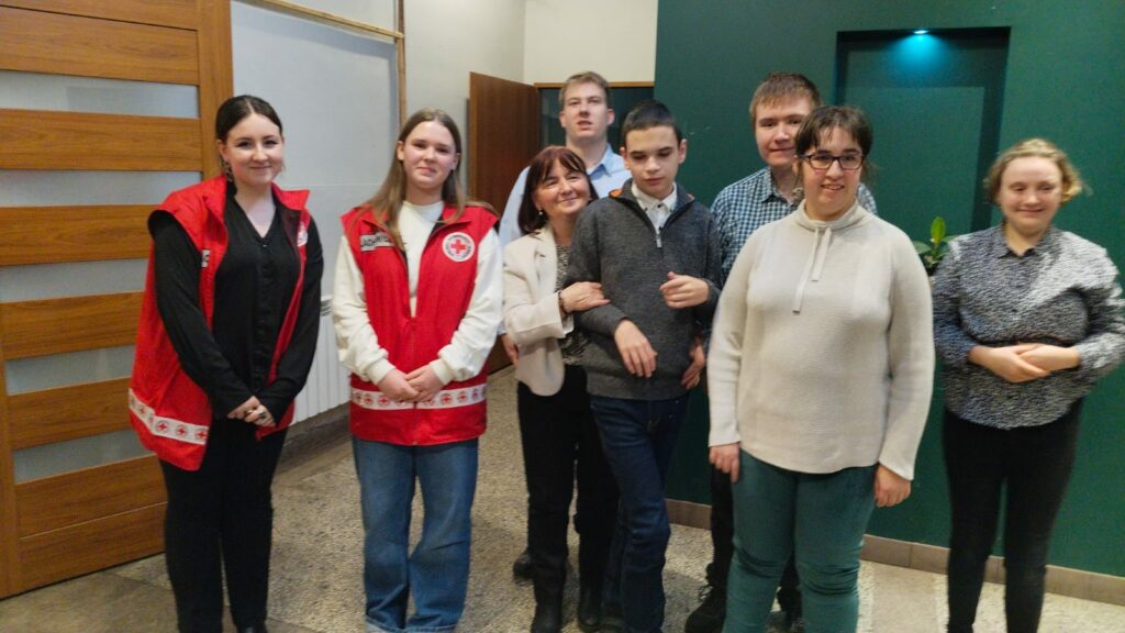 Grupa młodzieży pozuje do zdjęcia. Dwie młode kobiety mają założone czerwone kamizelki PCK.