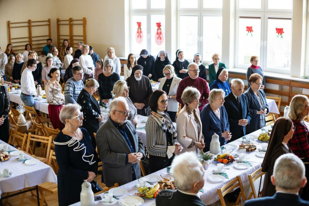 Grupa osób stoi przy stołach w jasnej sali, wspólna chwila skupienia podczas spotkania.