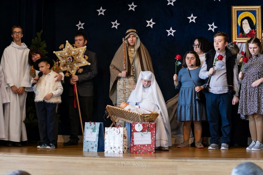 Grupa dzieci i dorosłych na scenie przedstawia jasełka - Maryja z Dzieciątkiem w żłóbku, Józef, kolędnicy i gwiazda betlejemska na tle gwiazd.