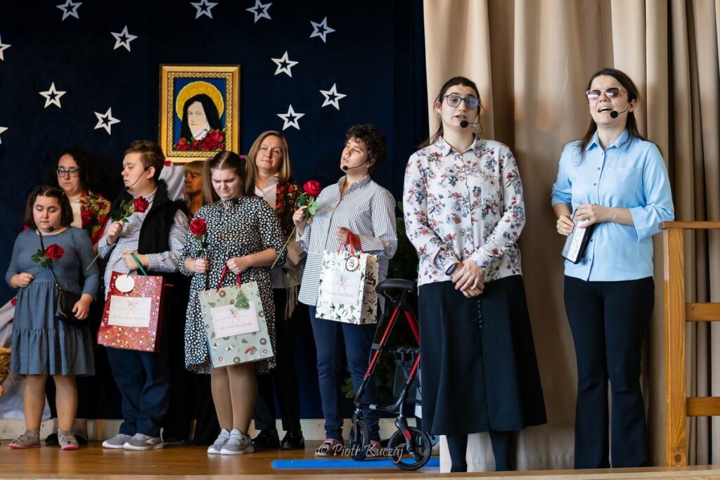 Aktorzy stoją na scenie trzymając róże i prezenty w rękach, a z prawej strony dwie młode kobiety śpiewają do mikrofonów.