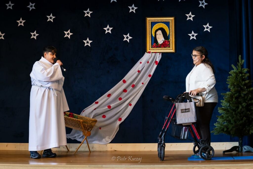 Scena przedstawienia – osoba w stroju anioła mówi do mikrofonu, a naprzeciw stoi młoda kobieta poruszająca się o balkoniku.