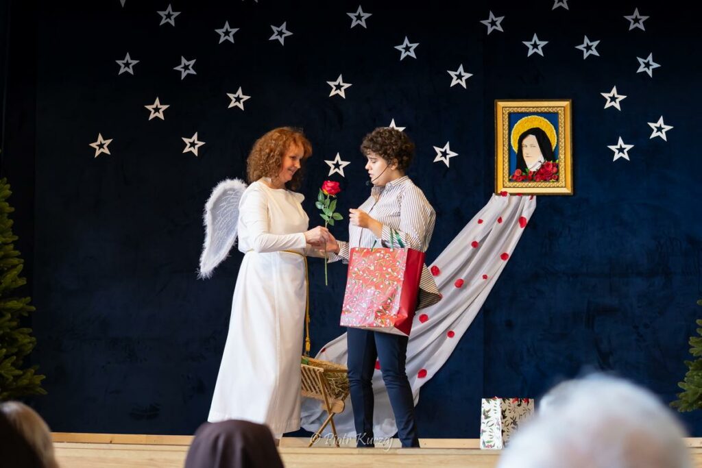 Scena przedstawienia – osoba w stroju anioła wręcza różę kobiecie trzymającej prezent, dekoracyjne tło z gwiazdami.