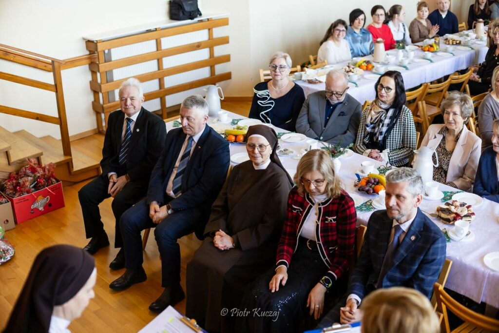 Uczestnicy uroczystego spotkania siedzą przy długich stołach nakrytych do poczęstunku w sali.