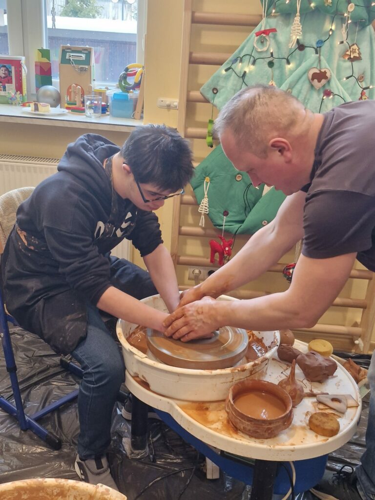 Chłopiec pracuje na kole garncarskim, a dorosły instruktor pomaga mu formować glinę.