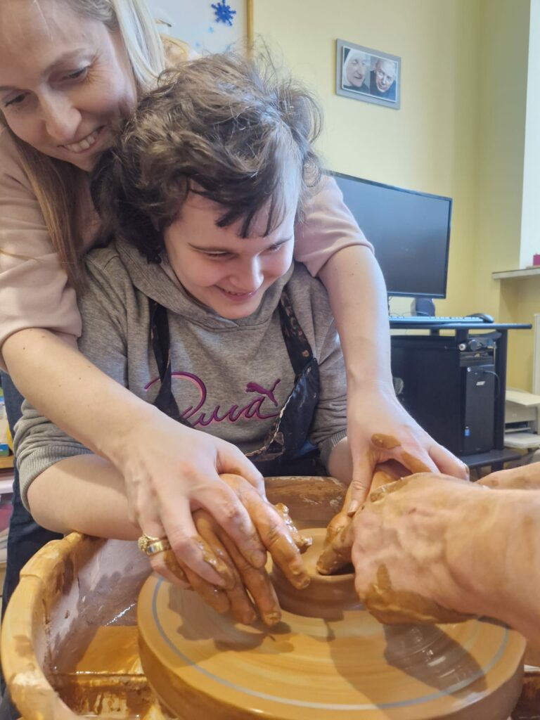 Uśmiechnięta dziewczynka pracuje na kole garncarskim z pomocą nauczycielki i instruktora.