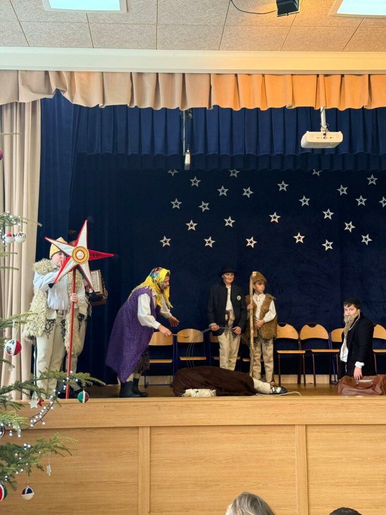 Dzieci w ludowych strojach stoją na scenie z dekoracją gwiazd.