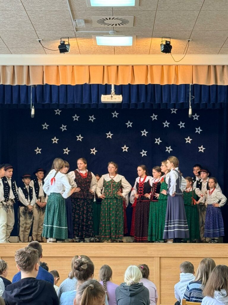 Dzieci w tradycyjnych strojach ustawione w rzędzie na scenie z gwiazdami w tle.