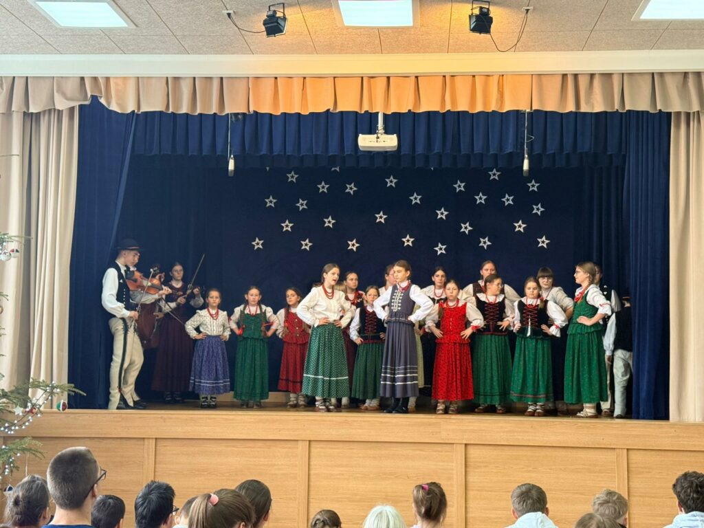 Grupa dzieci i młodzieży w strojach regionalnych stoi na scenie. po lewej stronie osoby grające na skrzypcach, w tle gwiazdy na niebieskim tle.