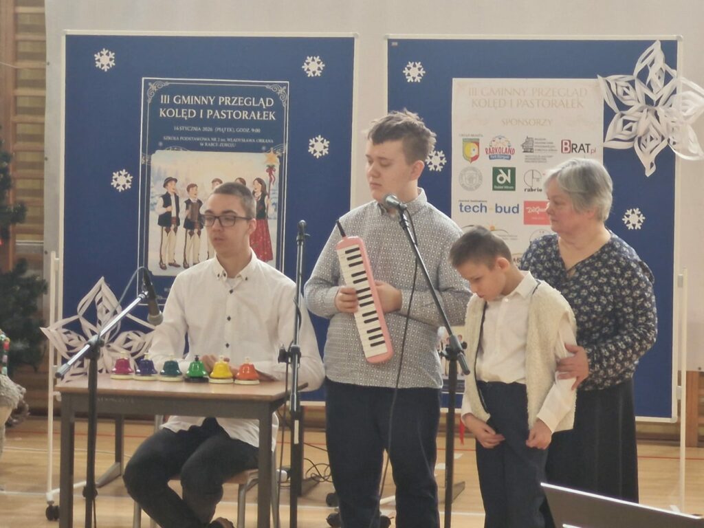 Trzech chłopców. Jeden siedzi przy stole i gra na dzwonkach naciskowych. Drugi ma w rękach melodykę i śpiewa do mikrofonu. Najmłodszy chłopiec stoi podtrzymywany przez dorosłą kobietę. 