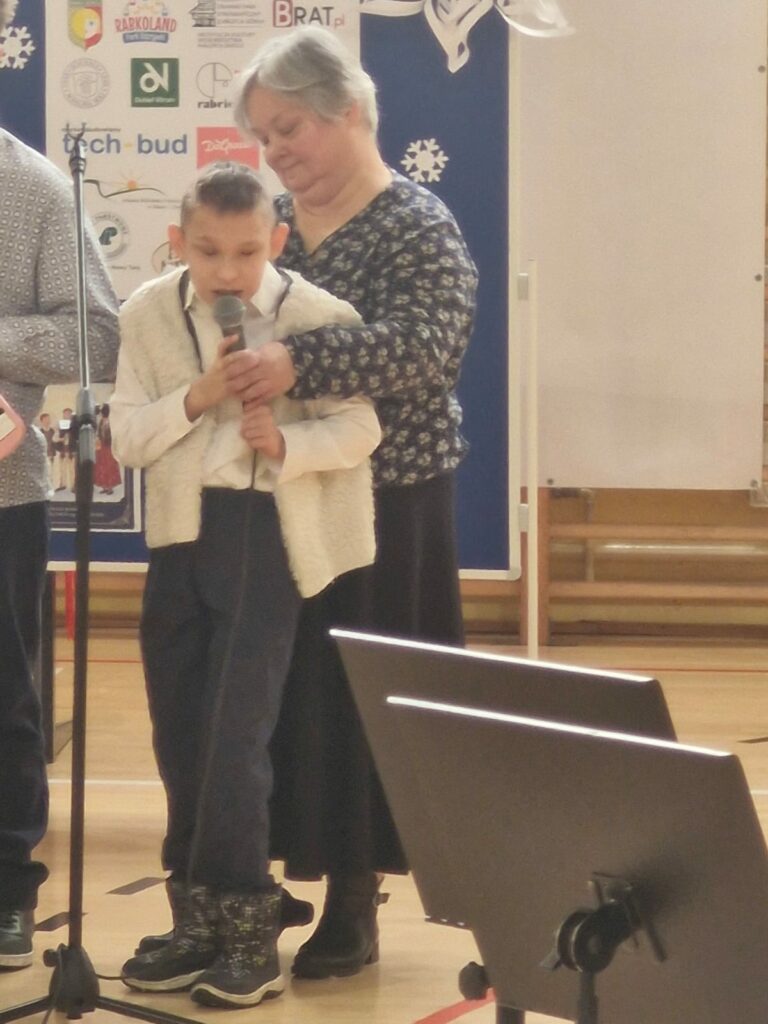 Chłopiec śpiewa do mikrofonu, który pomaga mu trzymać dorosła osoba.