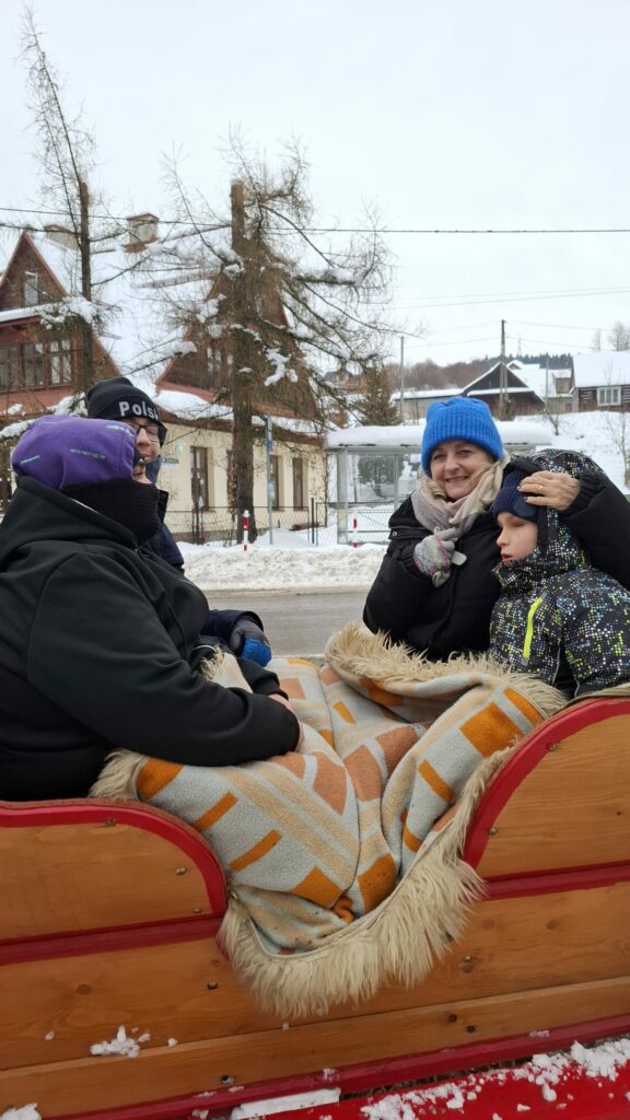 Grupa dorosłych i dziecko siedzą w saniach, owinięci kocami, podczas zimowej przejażdżki przez zaśnieżoną miejscowość.