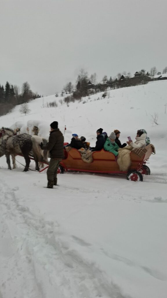 Grupa dorosłych i dzieci siedzi w saniach. obok znajduje się mężczyzna powożący. Sanie są zaprzężone w dwa konie.