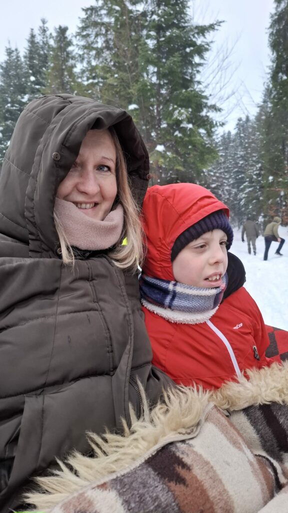 Kobieta i dziecko w ciepłych kurtkach siedzą w saniach, uśmiechnięci, na tle ośnieżonego lasu.
