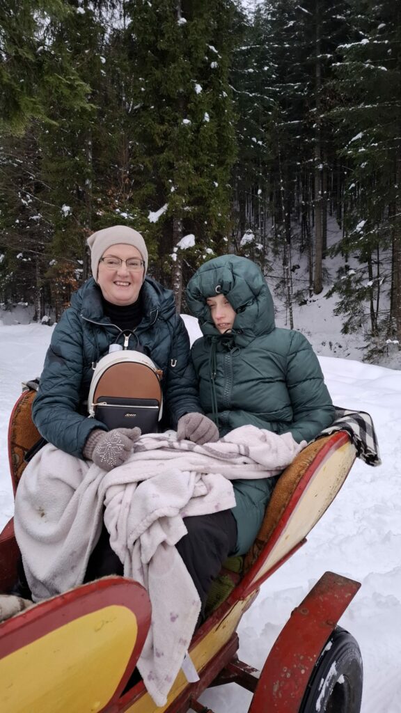 Dwie osoby siedzą w saniach, otulone kocem, w zimowym lesie.