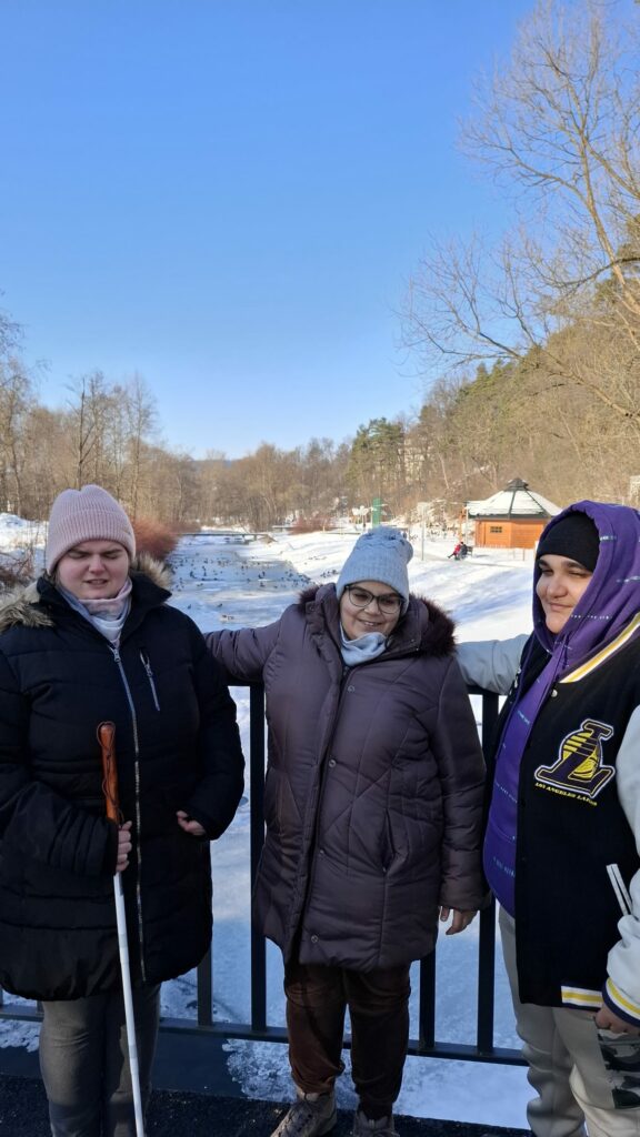 Dwie młode kobiety i mężczyzna stoją na mostku w tle widoczne bulwary oraz rzeka i pływające kaczki.