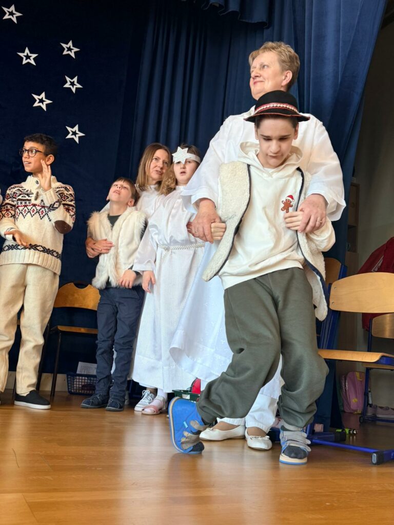 Dorośli z grupą dzieci stoją na scenie w strojach pastuszków i aniołów.