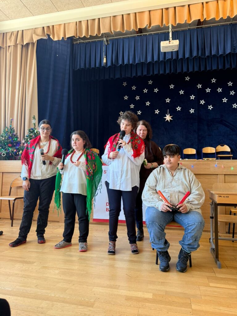 Grupa młodzieży w strojach regionalnych stoi pod sceną i śpiewa do mikrofonów, a chłopiec gra na dzwonkach.