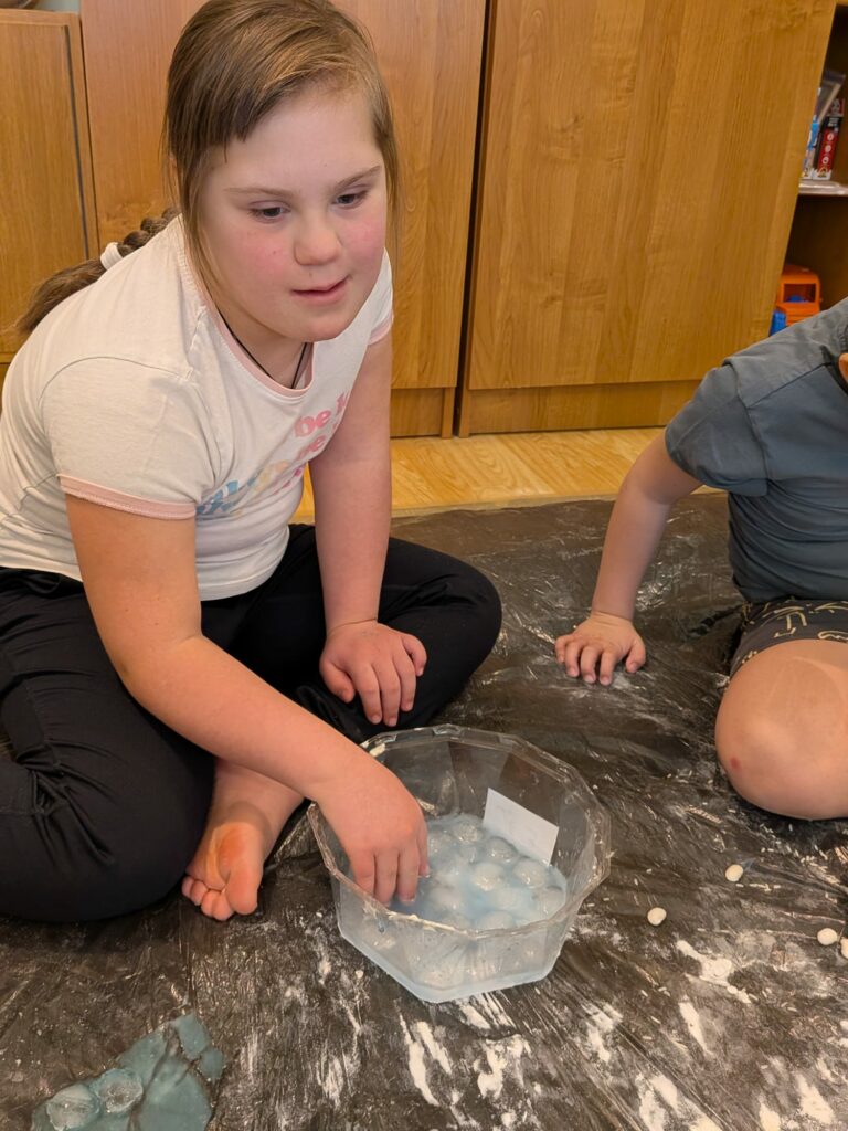 Dziewczynka siedzi na podłodze zabezpieczonej folią i wkłada dłoń do przeźroczystej miski wypełnionej niebieskawa cieczą i kostkami lodu.