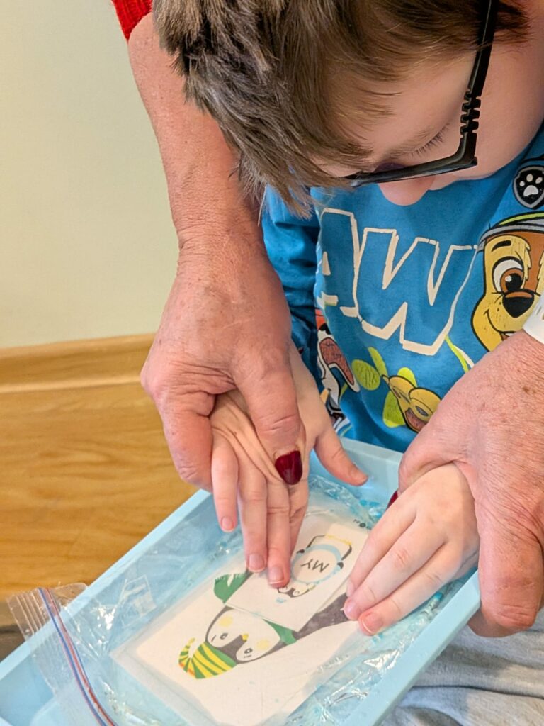 Dorosła osoba pomaga dziecku w okularach dotknąć karty z napisem "my" umieszczonej w niebieskiej tacy wypełnionej masą sensoryczną.