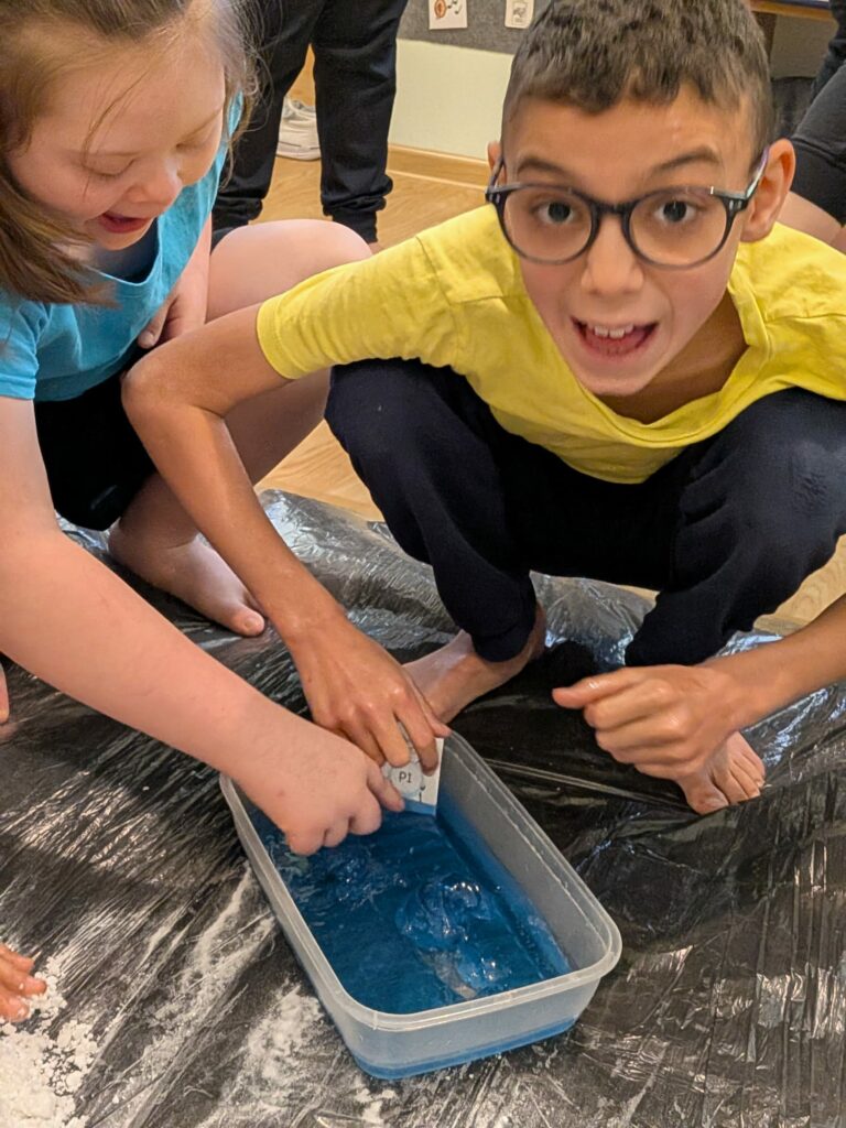 Dwójka dzieci bawi się przy pojemniku z niebieską cieczą . Jedno z nich zanurza w niej kartkę z napisem "pi".