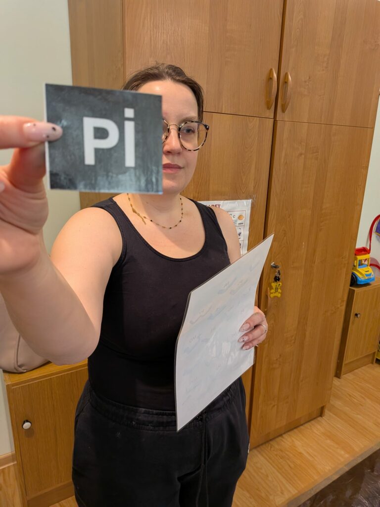 Kobieta w okularach trzyma przed twarzą małą kartkę z napisem "pi". W drugiej ręce trzyma zalaminowaną kartę z ćwiczeniami.
