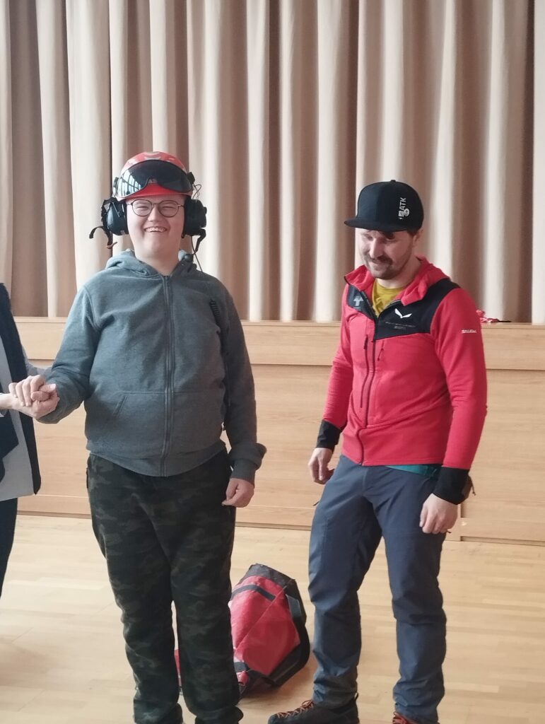 Trzy osoby stoją w sali; jedna z nich w kasku i słuchawkach trzyma nauczycielkę za rękę, obok leży sprzęt wspinaczkowy