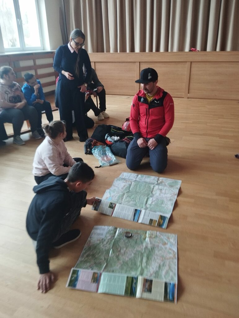 Instruktor klęczy na podłodze i pokazuje mapy grupie dzieci siedzących wokół sali.