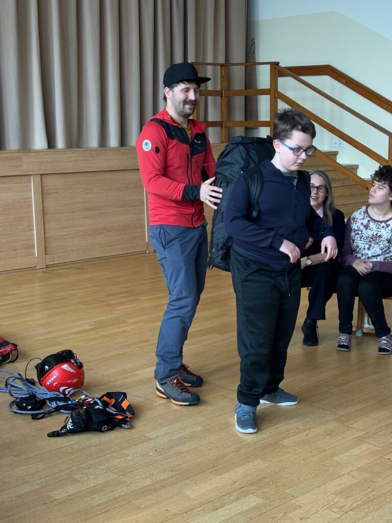 Mężczyzna w czerwonej kurtce pomaga chłopcu z plecakiem podczas zajęć w sali; w tle siedzą inni uczestnicy. 