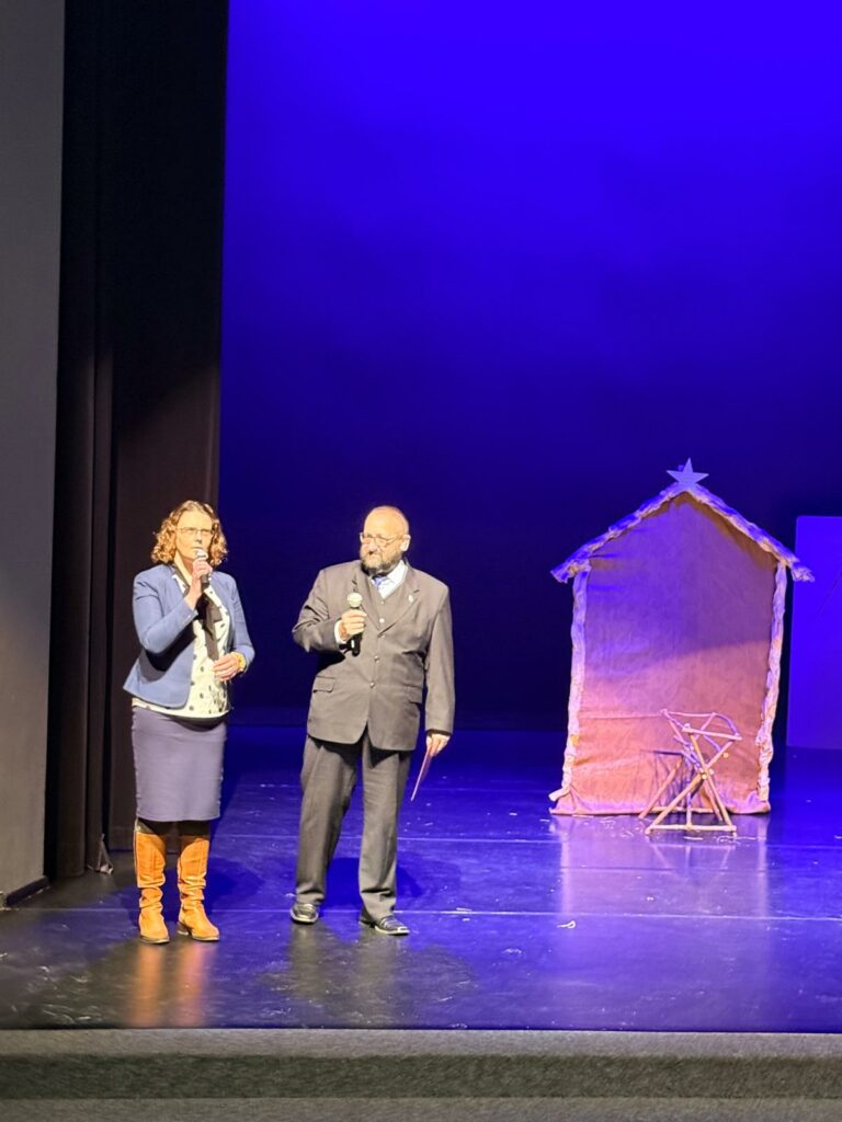 Dwie osoby dorosłe stoją na scenie teatralnej z mikrofonami, obok dekoracja szopki, całość oświetlona niebieskim światłem.