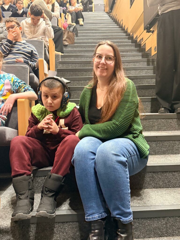 Kobieta siedzi na schodach sali obok chłopca w słuchawkach wygłuszających; oboje uczestniczą w wydarzeniu.