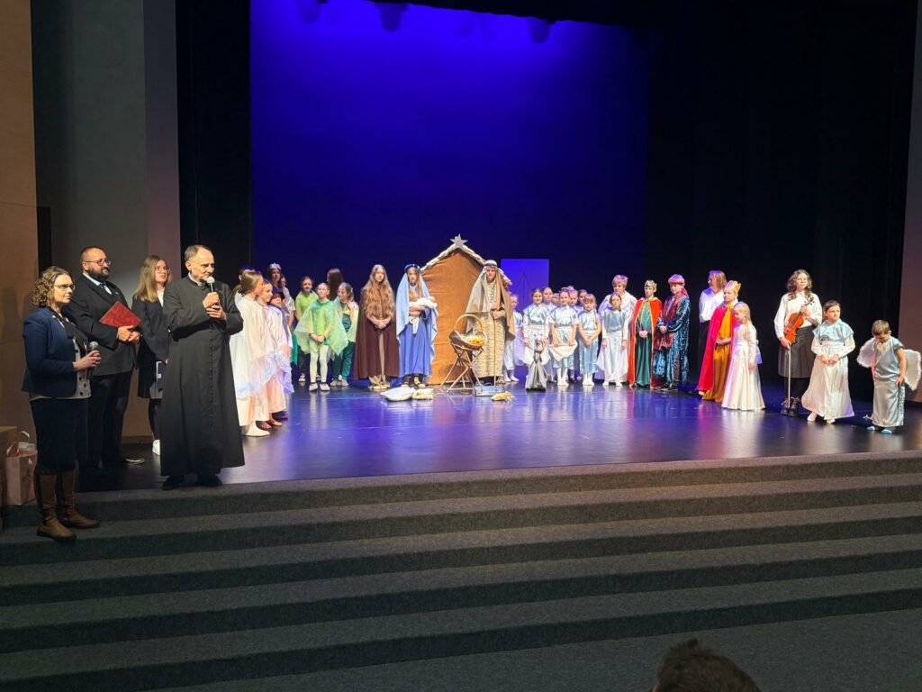 Duża grupa dzieci i dorosłych w strojach jasełkowych stoi na scenie wokół szopki, po zakończeniu przedstawienia.