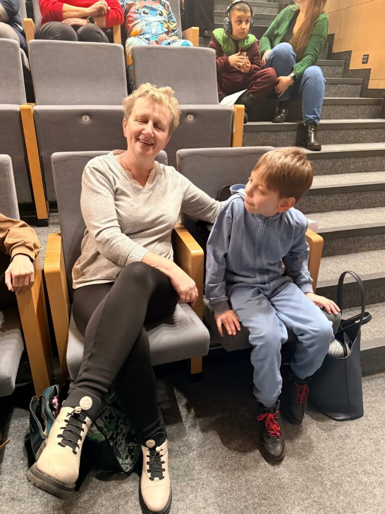 Kobieta siedzi na widowni obok chłopca w niebieskim stroju, uśmiechają sie podczas wydarzenia.