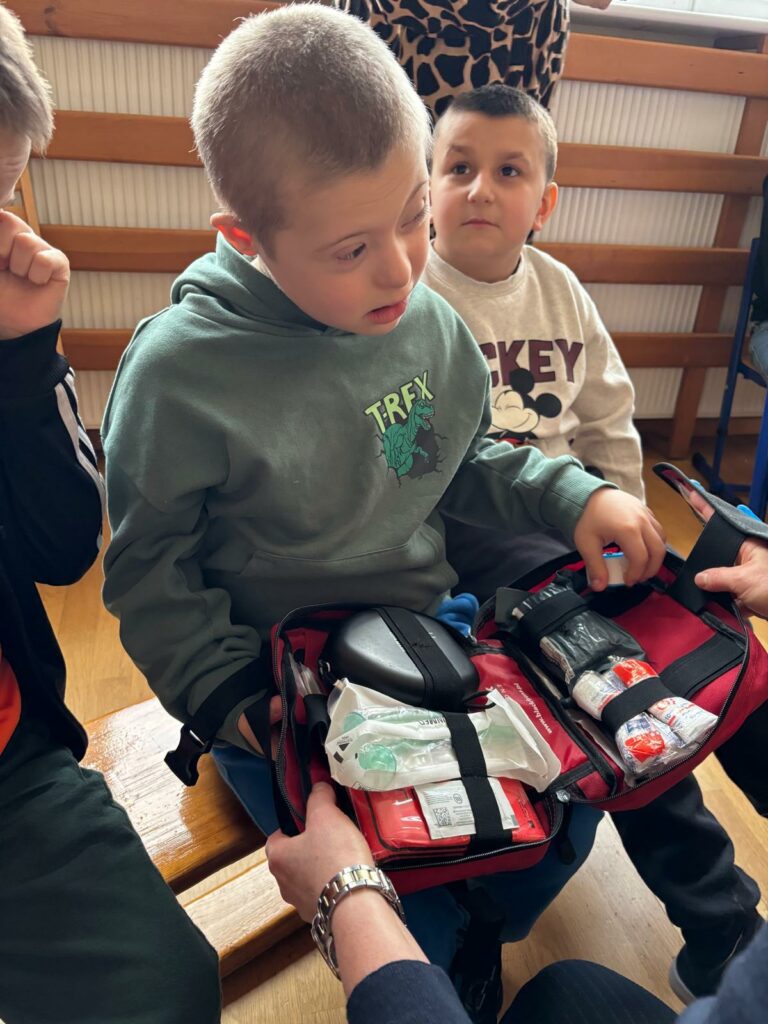 Dziecko siedzi na ławce i ogląda otwartą apteczkę pierwszej pomocy, obok inne dzieci obserwują z zainteresowaniem.