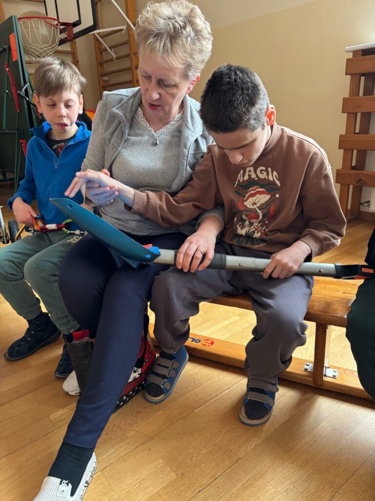 Kobieta i dwoje dzieci siedzą na gimnastycznej ławce. Kobieta pomaga chłopcu trzymać i oglądać niebieską łopatę.