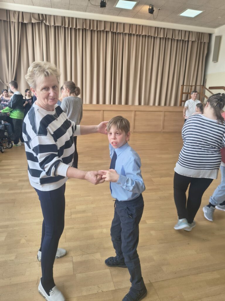 Nauczycielka tańczy z uśmiechniętym chłopcem w eleganckiej koszuli i krawacie podczas zabawy na szkolnej sali