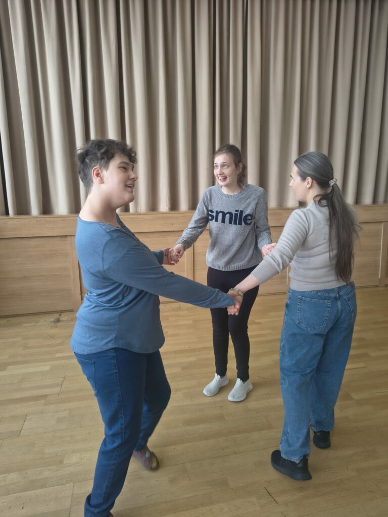 Dwie uczennice i nauczycielka trzymają się za ręce i tańczą w kręgu śmiejąc się.