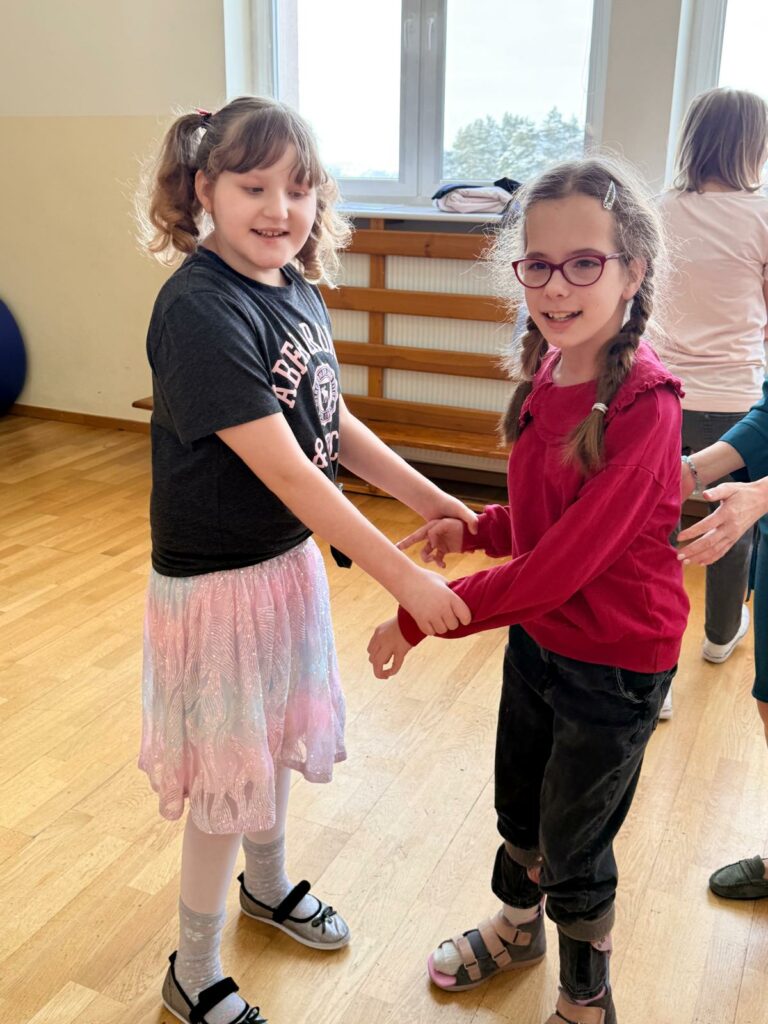 Dwie dziewczynki trzymają się za ręce podczas zabawy w jasnej sali z drewnianą podłogą.