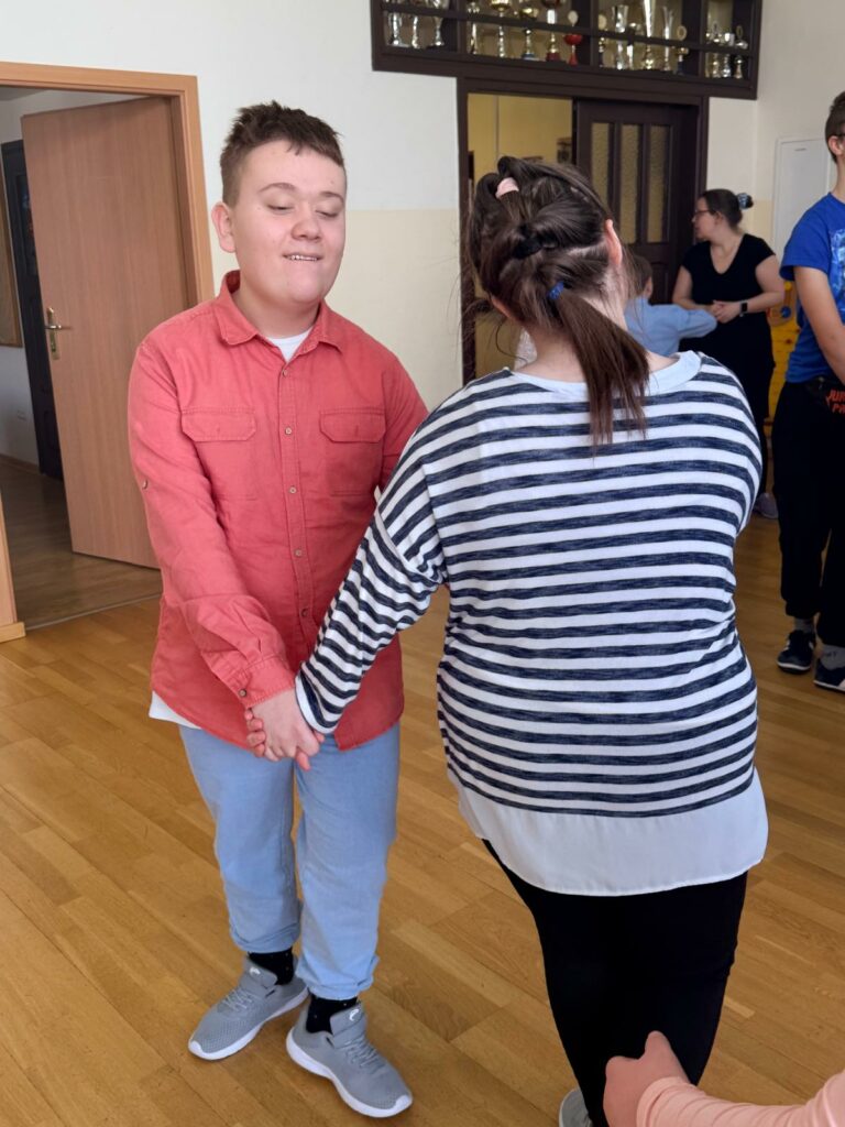 Dwójka osób trzyma się za ręce i tańczy, w tle widać inne osoby stojące przy drzwiach.